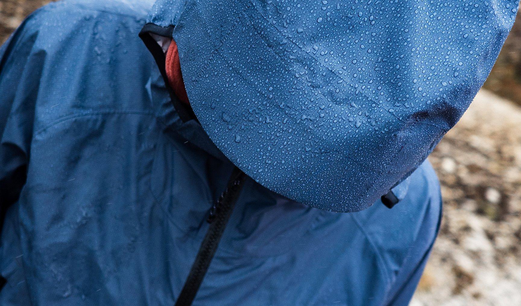 Close-up of a person wearing a dark blue Mountain Hardwear waterproof jacket and hood, both covered in raindrops. The individual is looking downward, emphasizing the water-repellent performance of the gear in wet, rugged outdoor conditions.