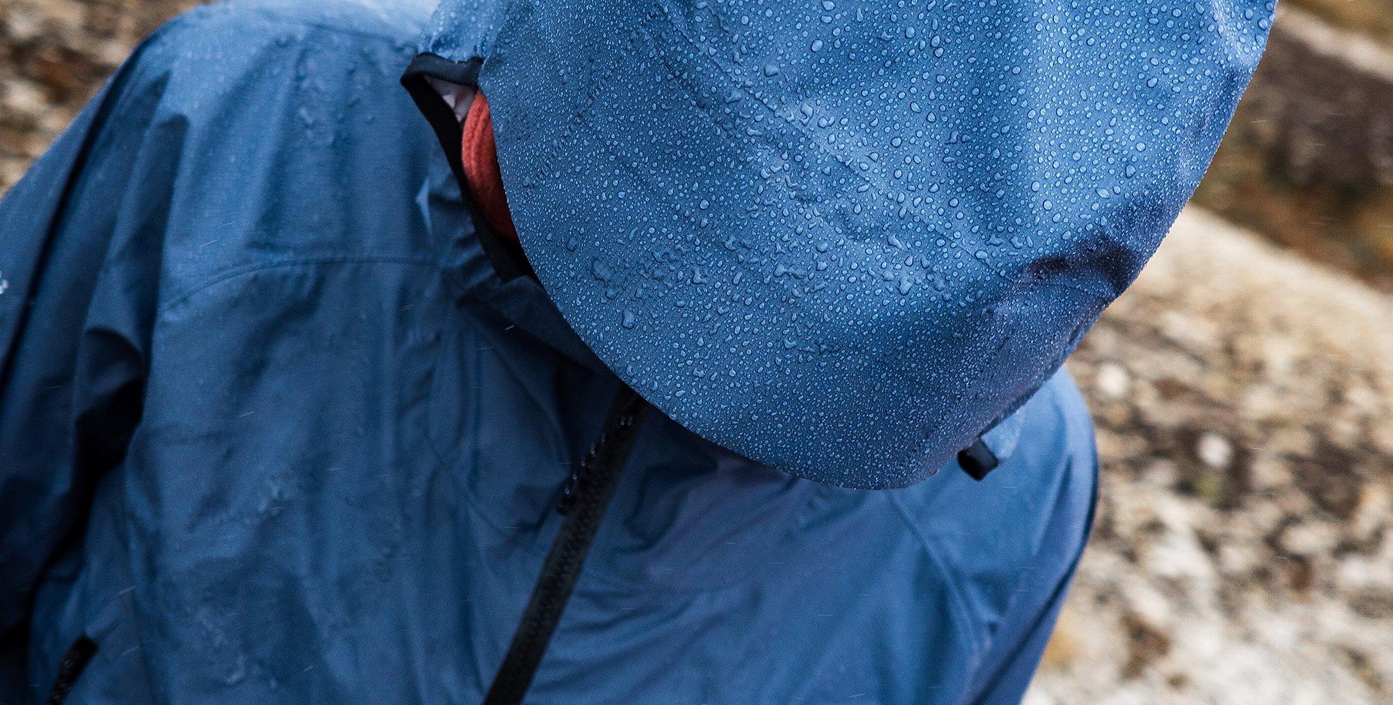 Close-up of a person wearing a dark blue Mountain Hardwear waterproof jacket and hood, both covered in raindrops. The individual is looking downward, emphasizing the water-repellent performance of the gear in wet, rugged outdoor conditions.