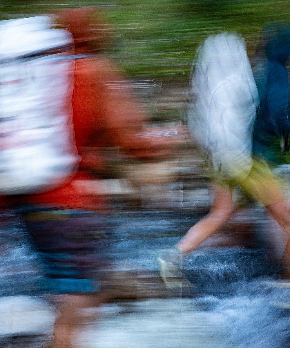 Abstract motion blur image of hikers crossing a stream, wearing large backpacks and outdoor gear. The photo captures dynamic movement with streaks of color, emphasizing speed, activity, and the energy of backcountry adventure.