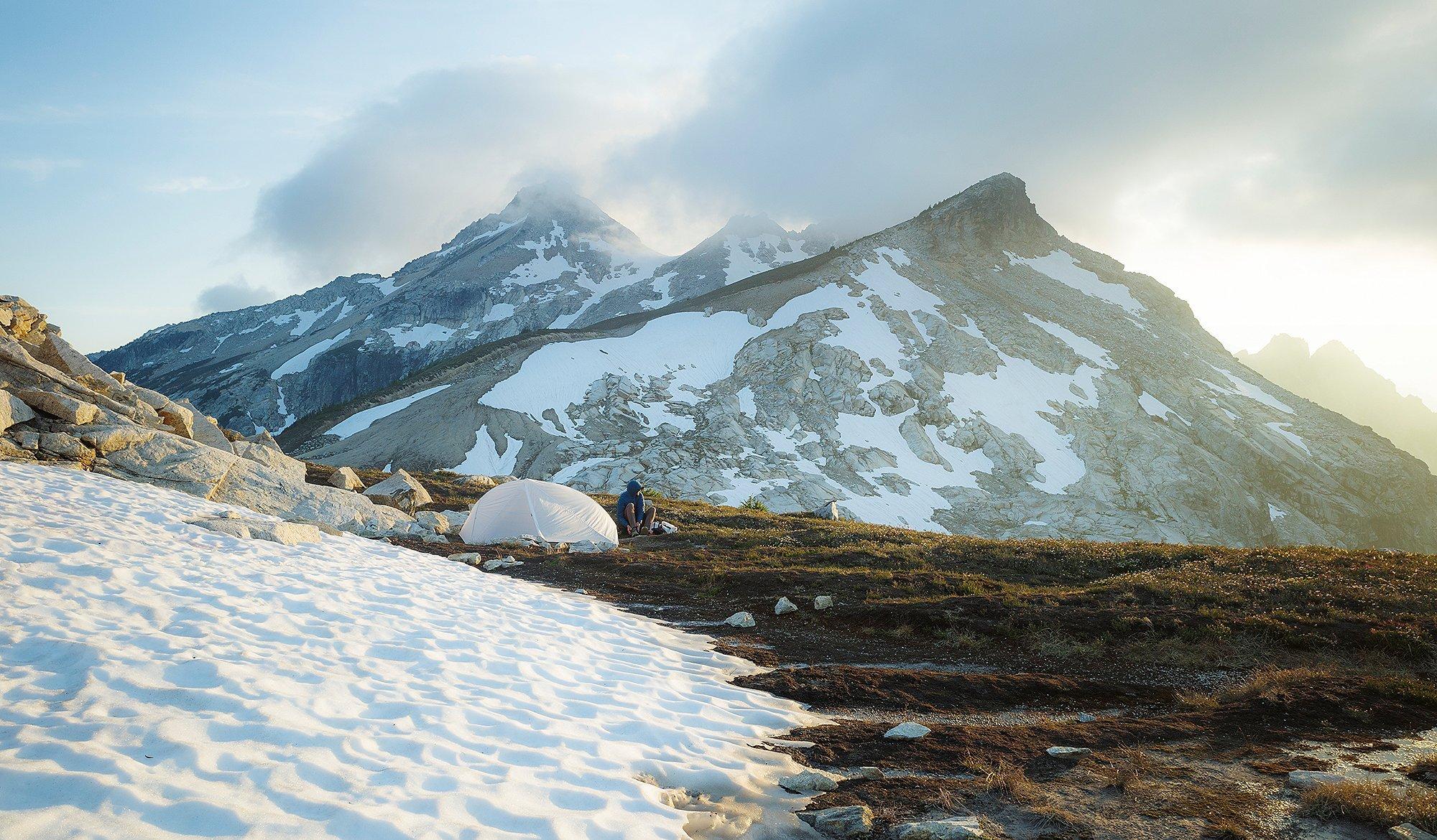 CJ sits beside a white MHW tent at sunrise, surrounded by alpine terrain with patches of snow and rugged granite peaks in the background. The scene captures a peaceful moment high in the mountains, with soft morning light breaking through clouds over the dramatic ridgeline.








