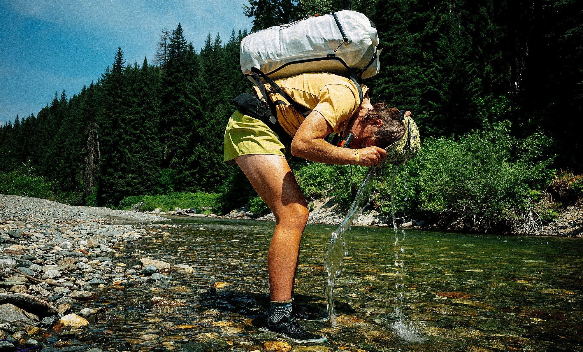 Hannah takes a sip of water from the river, hunched over with her pack.







