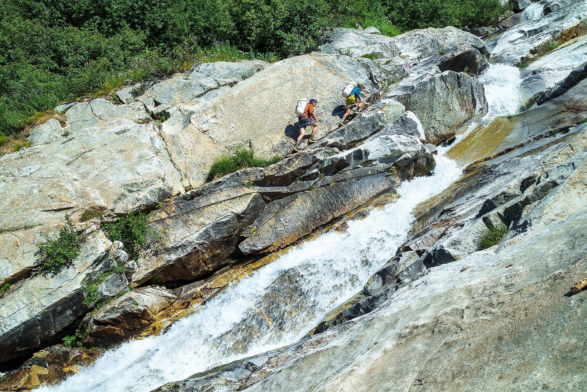 Two hikers with backpacks scramble up steep, sunlit granite slabs beside a rushing waterfall, surrounded by dense green forest at the top of the slope.