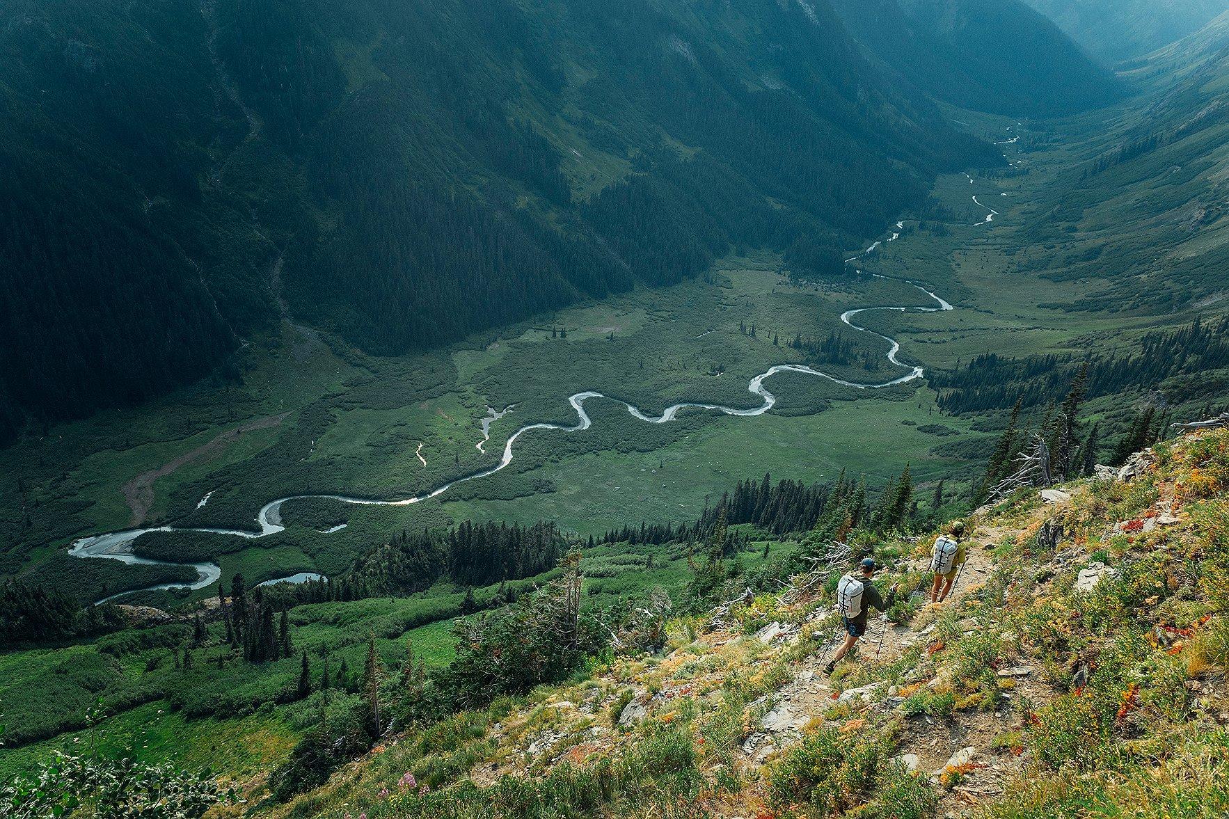 CJ and Hannah with backpacks and trekking poles descend a steep, flower-lined trail overlooking a vast, winding river that snakes through a lush, green valley surrounded by forested mountains.