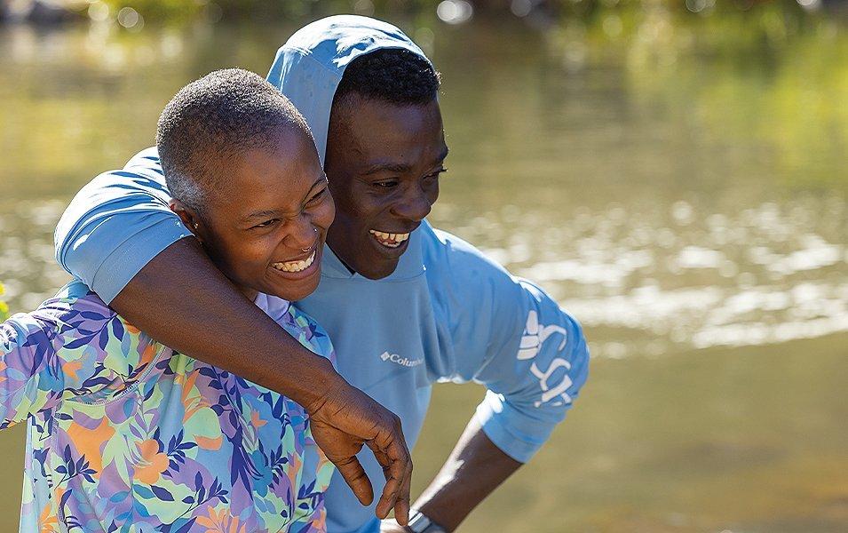 A man wearing an UPF shirt puts his arm around a kid and smiles with a scenic river in the background. 