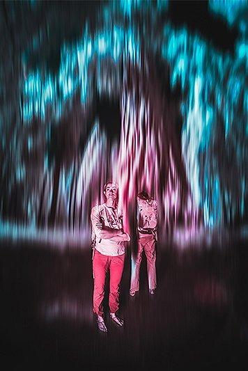Infrared image of two campers at night in the foreground, blue and pink light lines in the background. 
