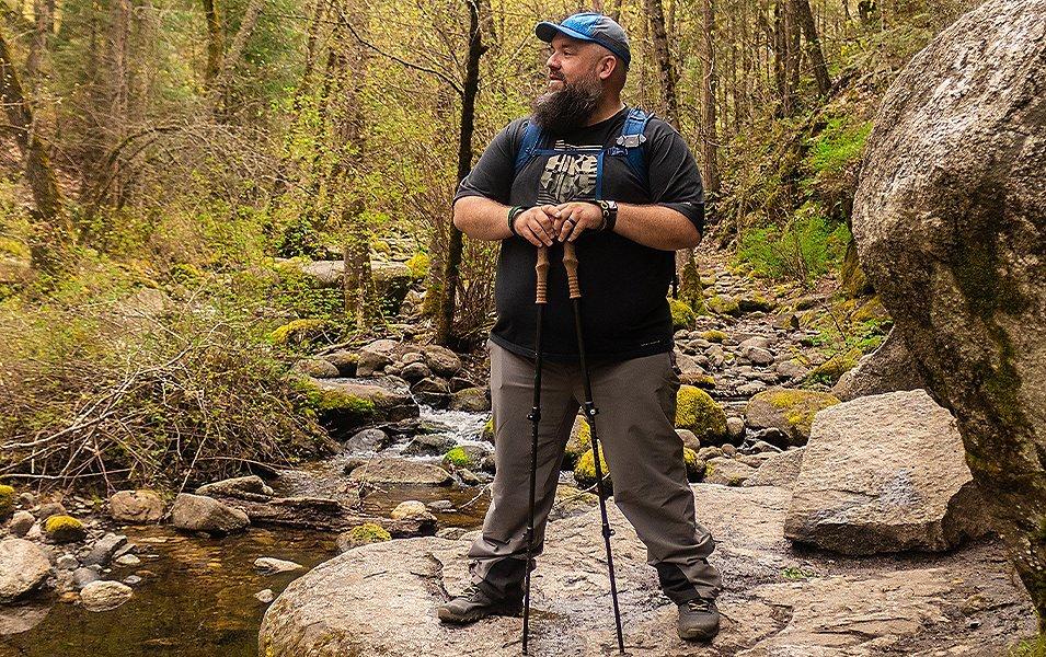 Andy Neal, an outdoor filmmaker and host of The Hiker Podcast, stands in a scenic wooded area holding a pair of hiking poles.