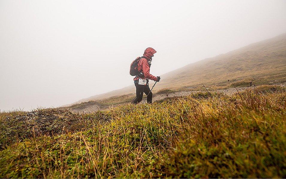 A man in a red jacket treks across a foggy hill holding hiking poles.