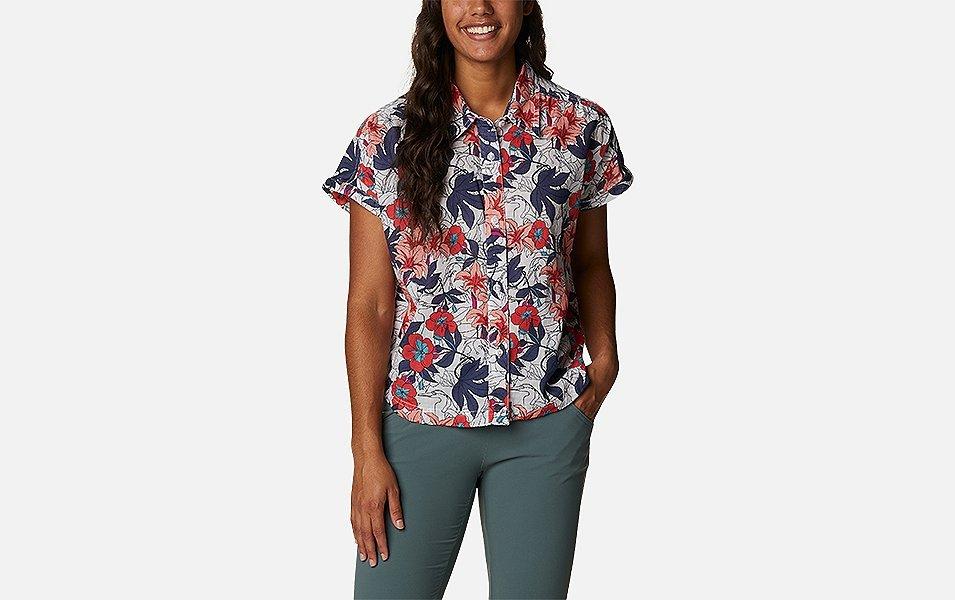 A product shot of a woman wearing a pink-and-purple floral Columbia Sportswear Camp Henry IV button-down shirt and blue pants with a gray background.