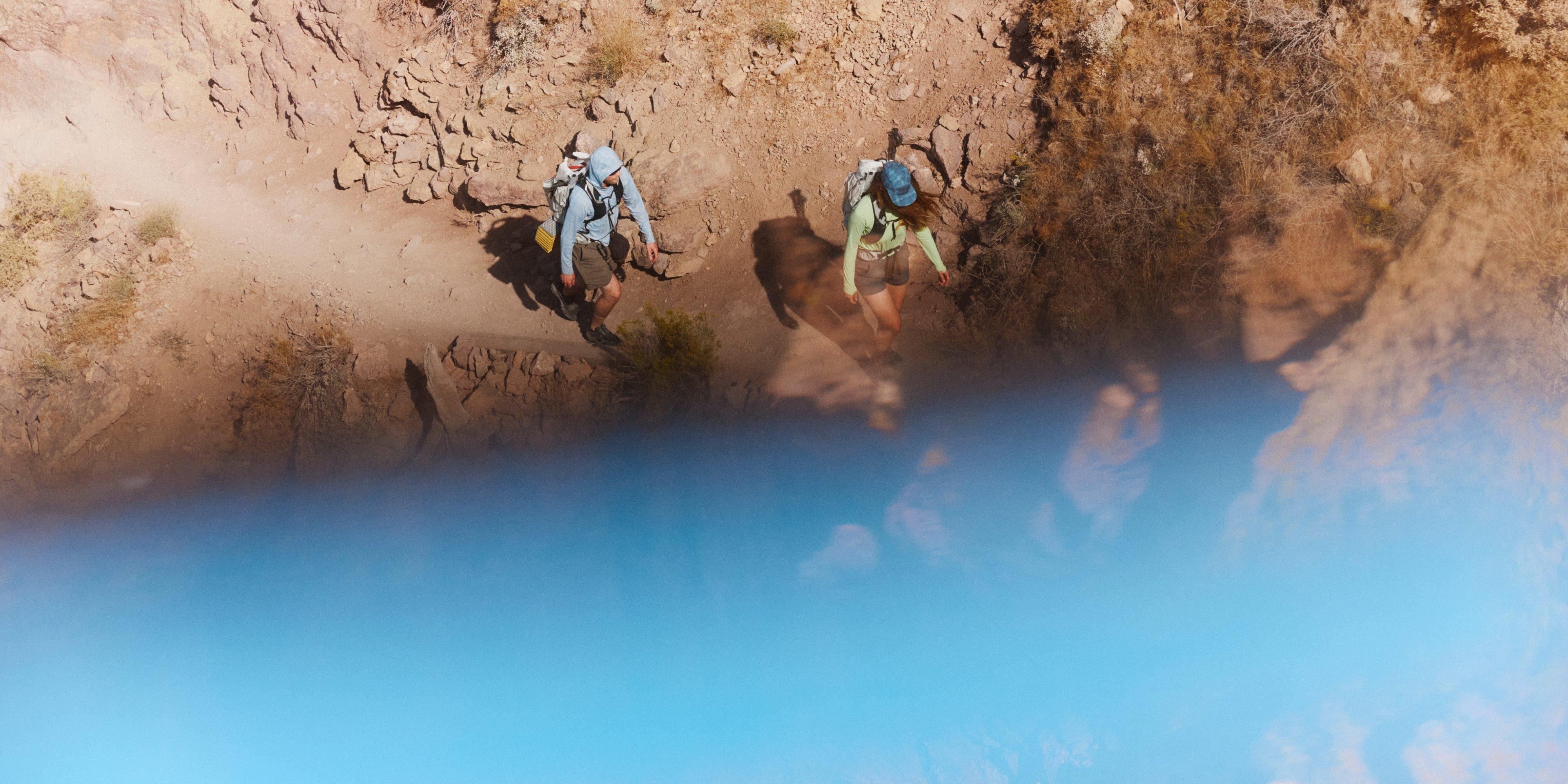 An aerial view of a man and woman in a Crater Lake™ Hoody and Long Sleeve, traversing a the barren dirt and rock path with no shade.