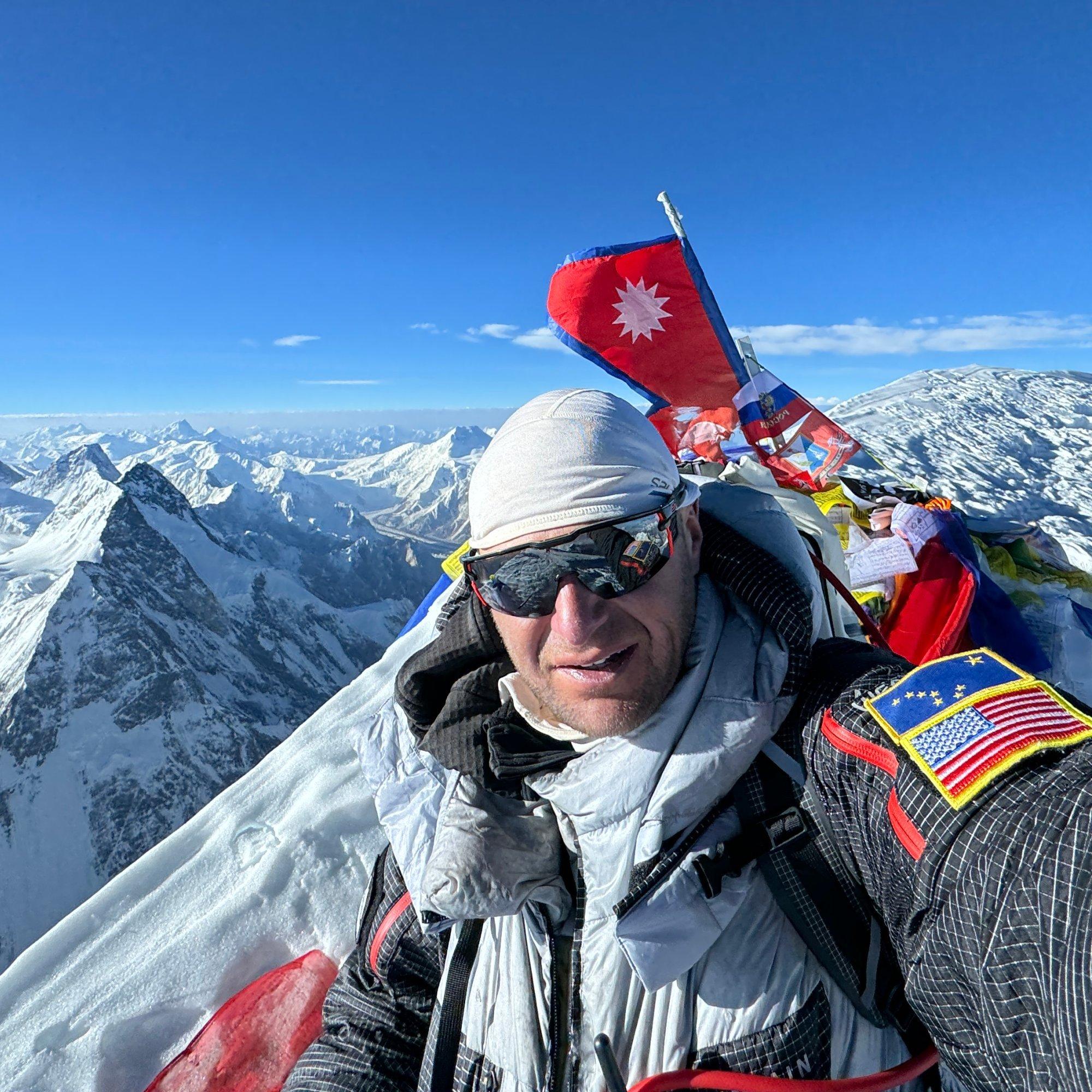 Professional alpine climber and mountaineer Benny Lieber takes a summit selfie, snowy peaks and prayer flags behind him.
