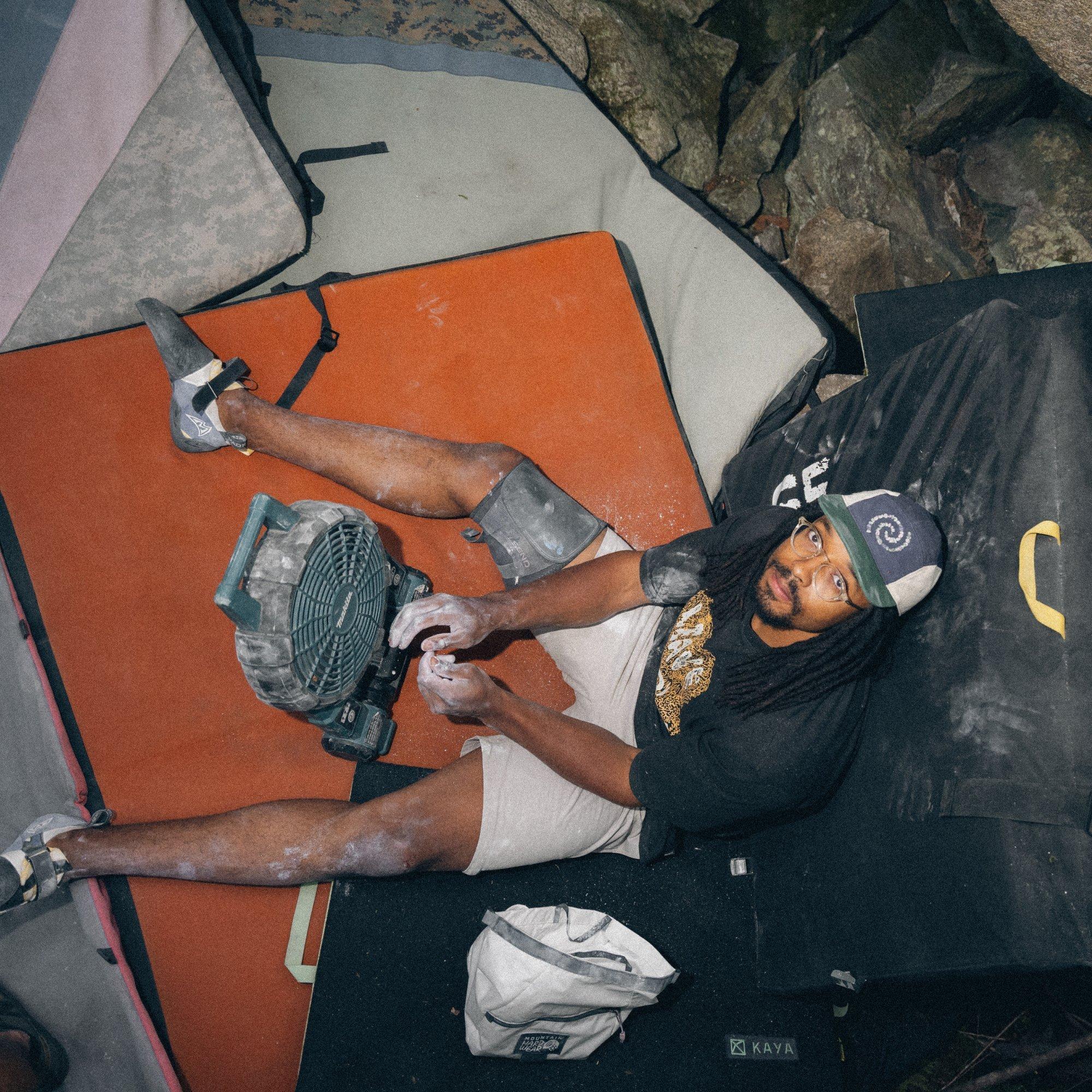 Professional outdoor climber Caleb Robinson looks up at the camera while seated on a crash mat, drying chalky hands in front of a fan.