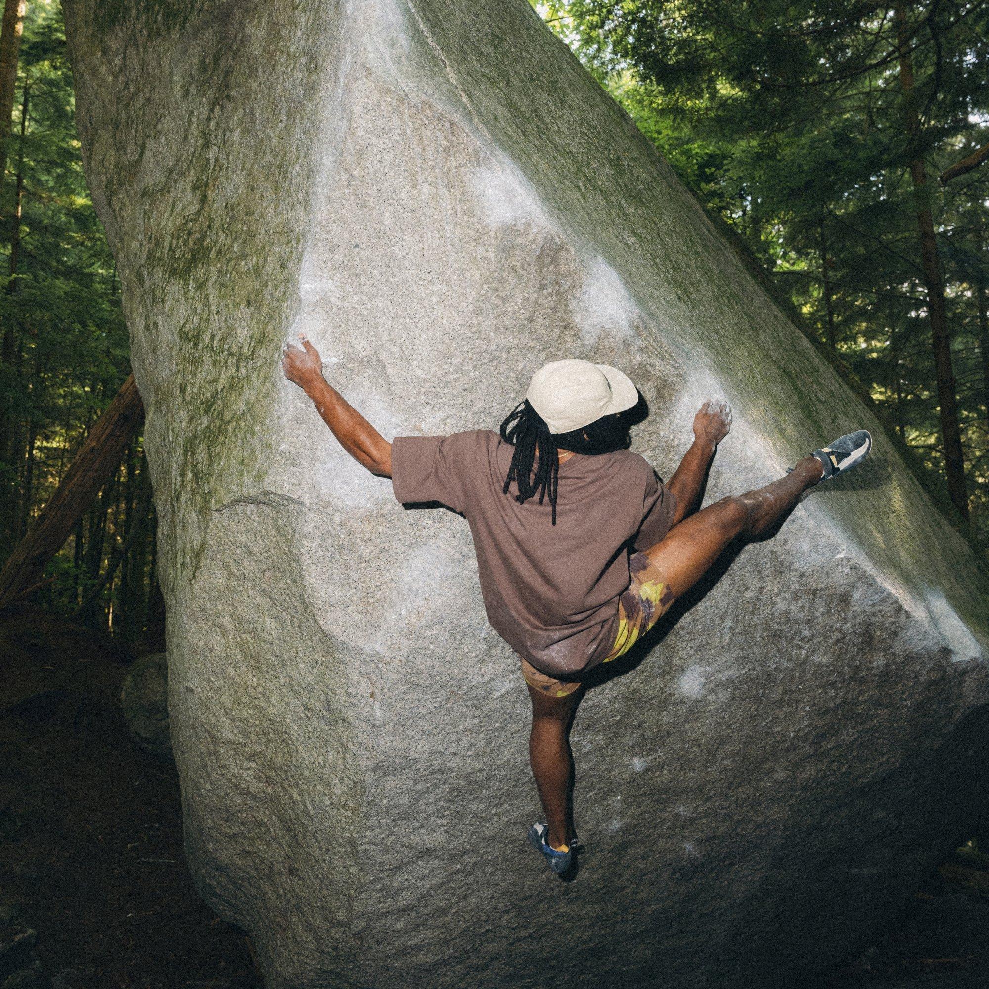 The back of his shirt dusty with chalk, professional outdoor climber Caleb Robinson reaches for a heel hook on a forest boulder.