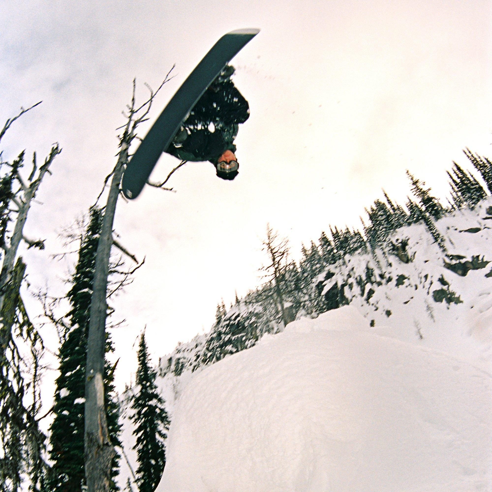 Completely inverted, professional snowboarder Emma Crosby soars through the air, a snowy tree line behind her.