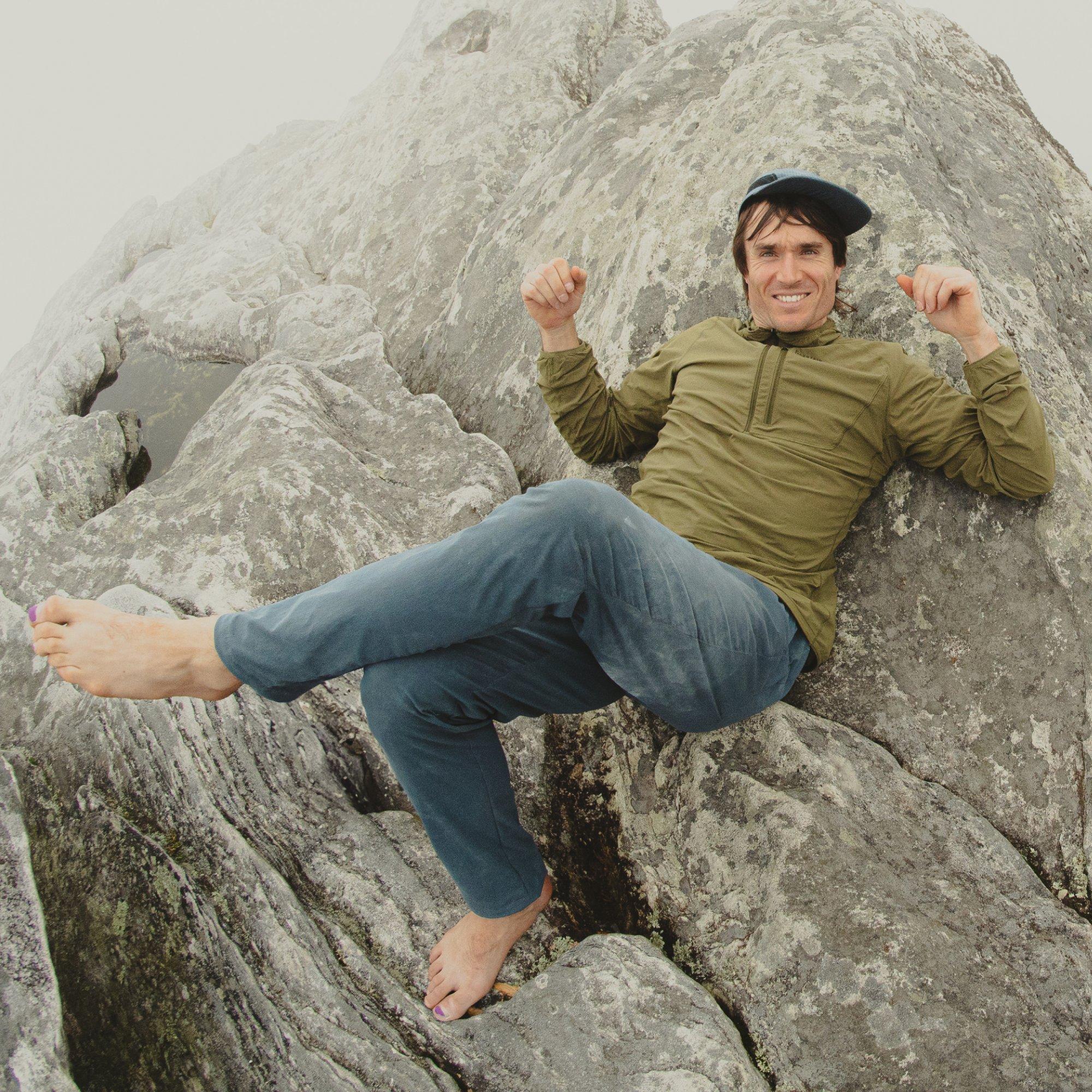 Professional climber Ethan Pringle reclines casually on a boulder cloaked in fog with bare feet, dusty pants, and a smile.