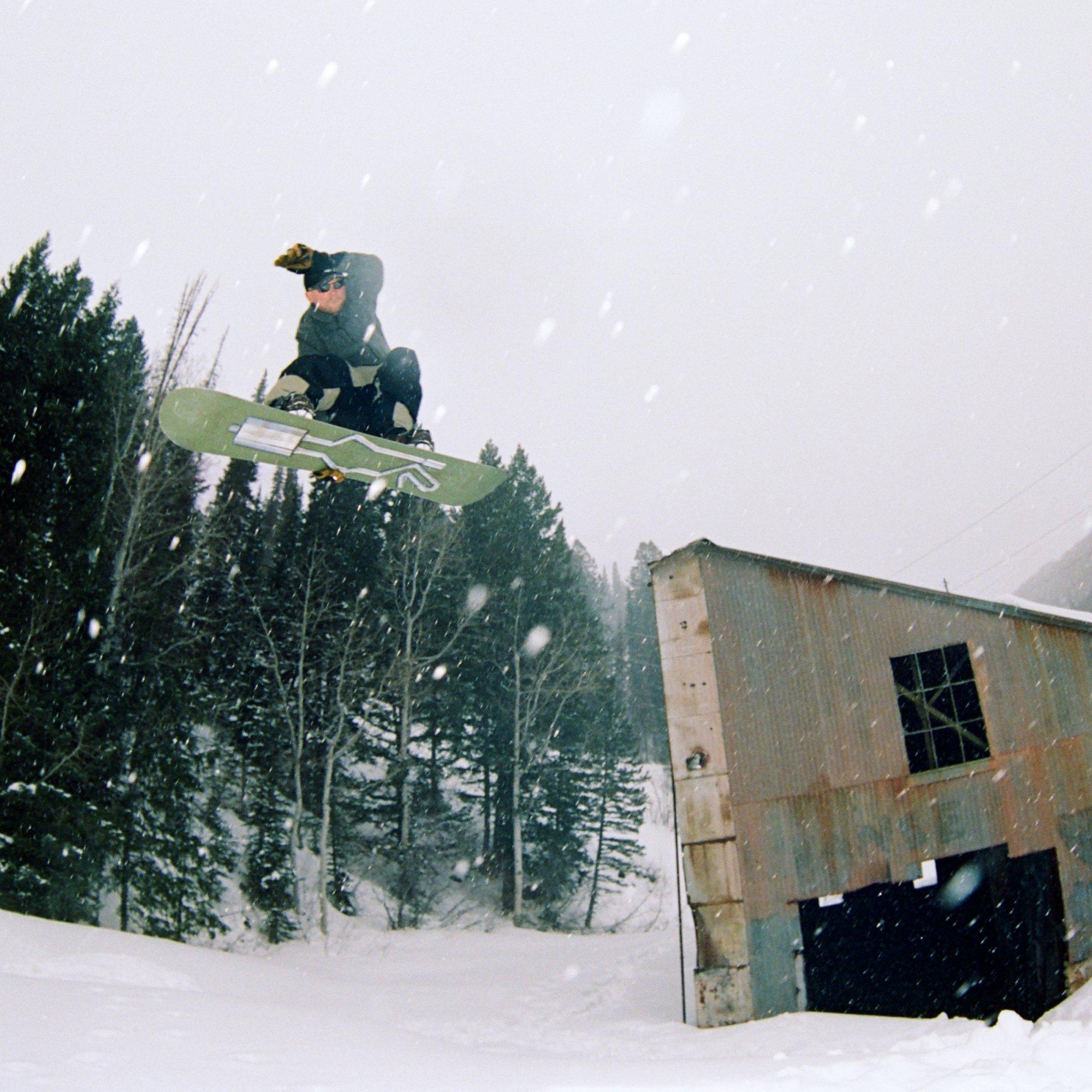 Professional snowboarder Jake Kuzyk launches off a jump, a decrepit barn and snowy trees in the background.