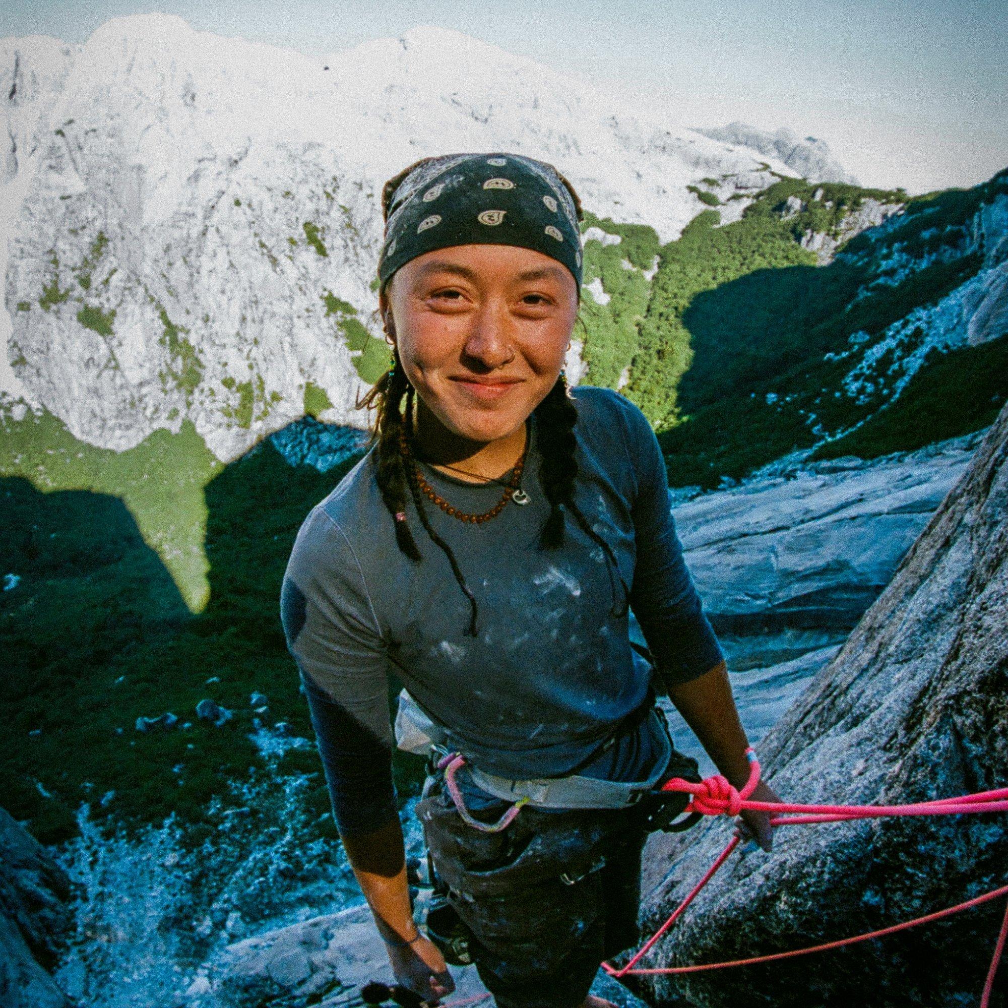 The valley floor visible below, professional climber Rell Lennox holds her line and offers a smile, paused barefoot on a rock ledge.