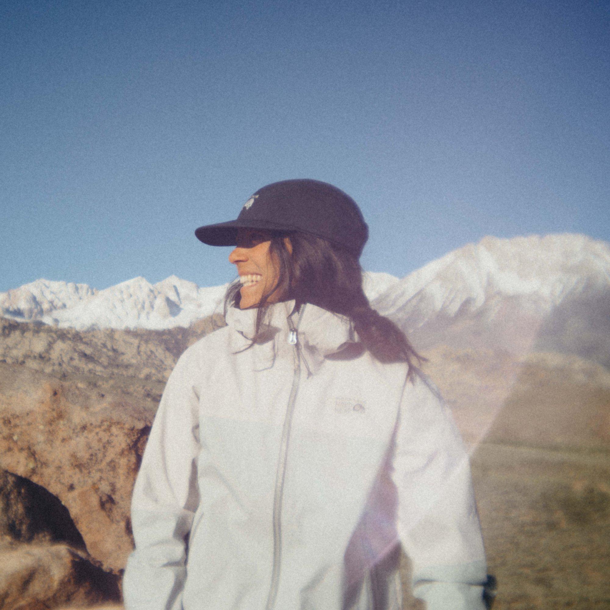 Grinning as she looks over her shoulder, professional trail athlete and photographer Sarah Attar poses in front of a mountain range.