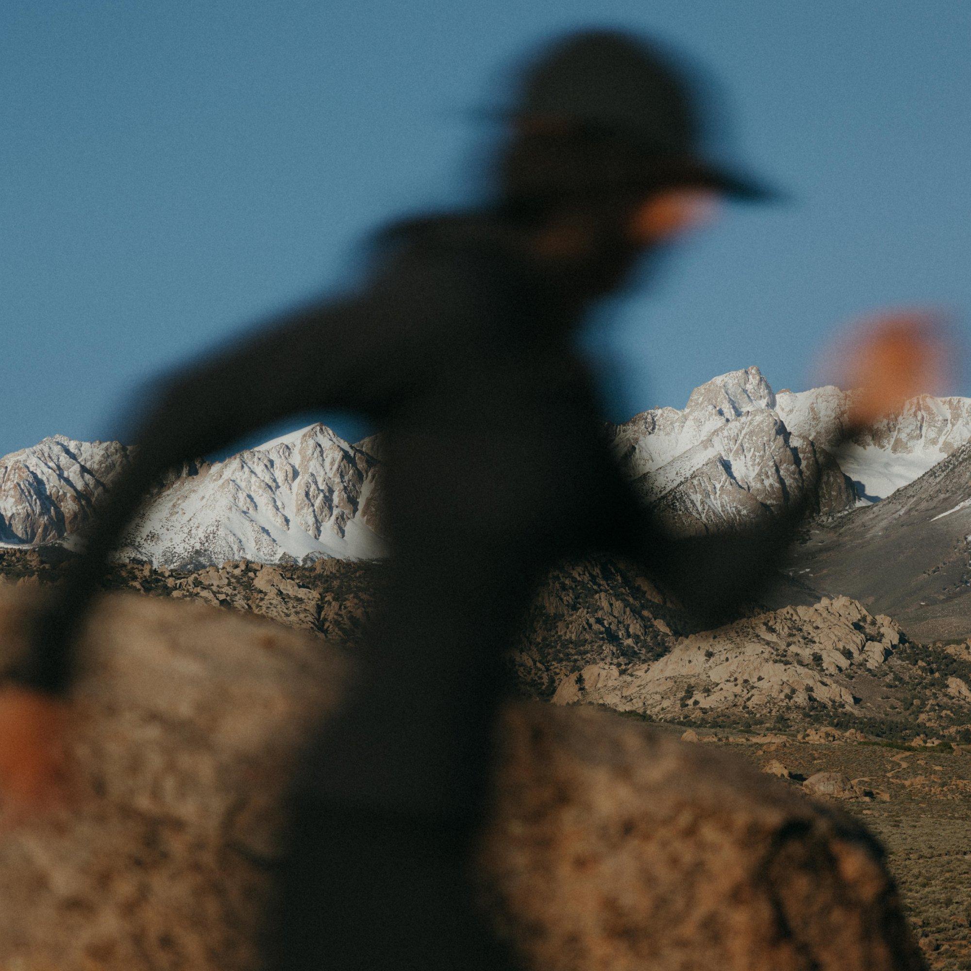The camera focused on the range of rocky mountains behind her, professional trail athlete Sarah Attar runs by, seen in silhouette.