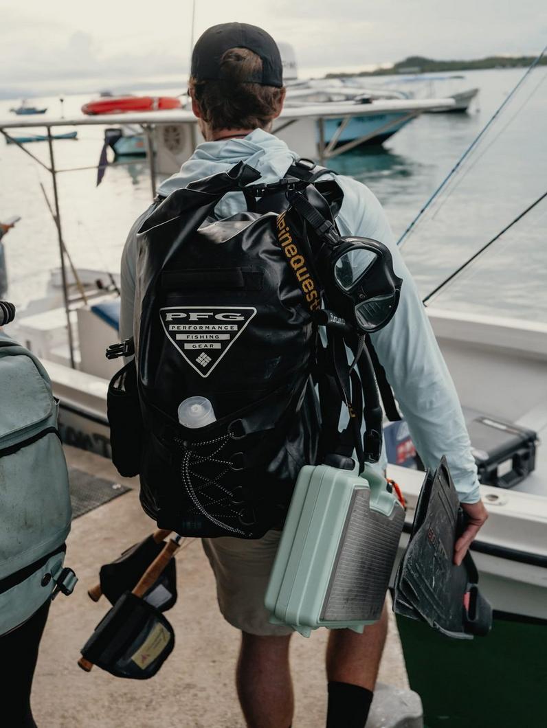 Person wearing black waterproof fishing backpack and carrying tackle box at marina dock.