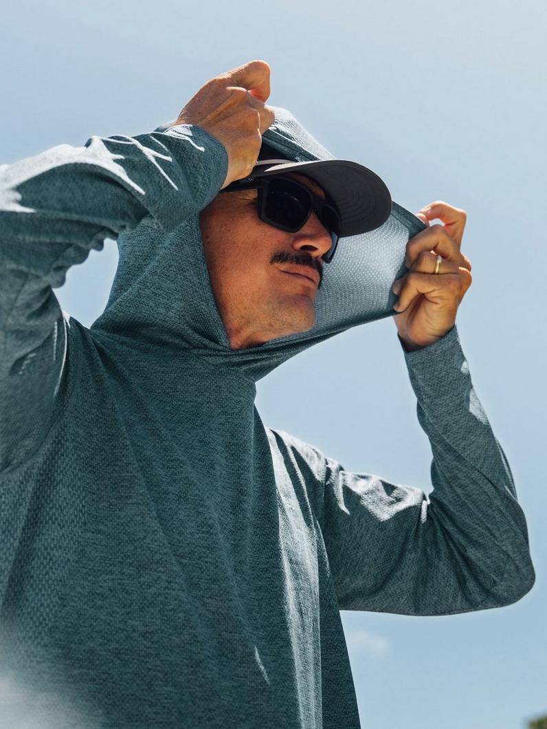 Man pulling on teal hoodie with white graphic design while wearing sunglasses and black cap.