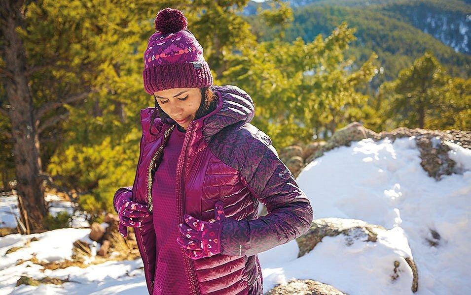 A woman wearing a fleece and puffer jacket stands on a scenic winter hiking trail looking down at the ground. 