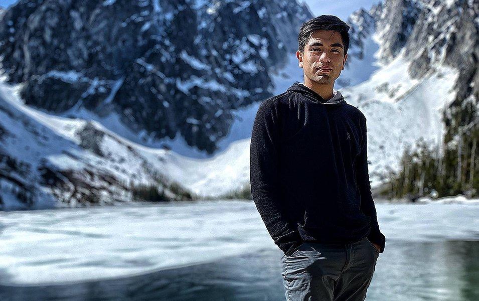 Allen Cárdenas stands with his hands in his pockets smiling at the camera as gorgeous mountains overlook an alpine lake in the background.