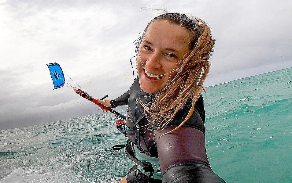 Pro kiteboarder Michaela Pilkenton smiles widely at the camera in a wetsuit as she soars across the water holding her kite in the background.