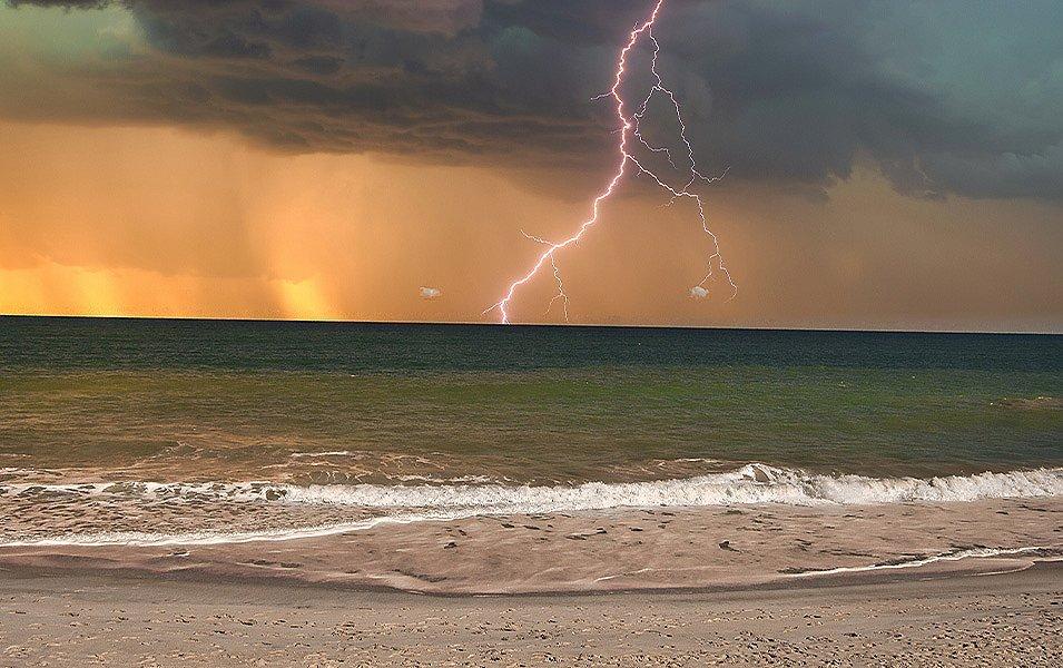 A big thunderstorm crashes down over an ocean. In the forefront are waves and a sandy beach; in the background, there are black clouds. A jagged bolt of lightning strikes the water as the sun sets to the west.  