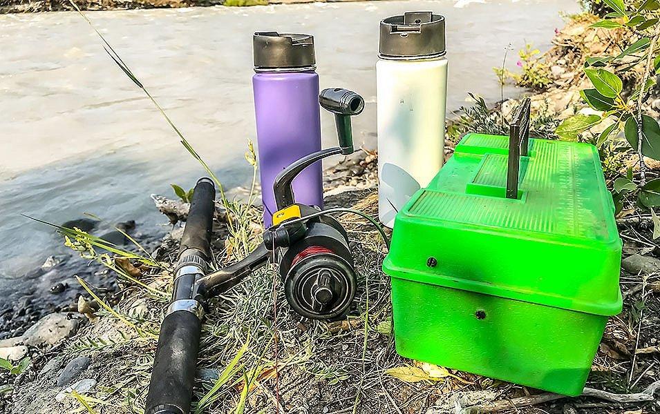 A close-up shot of a pile of fishing gear sitting by a river bank that includes a fishing rod, water bottles, and tackle box. 