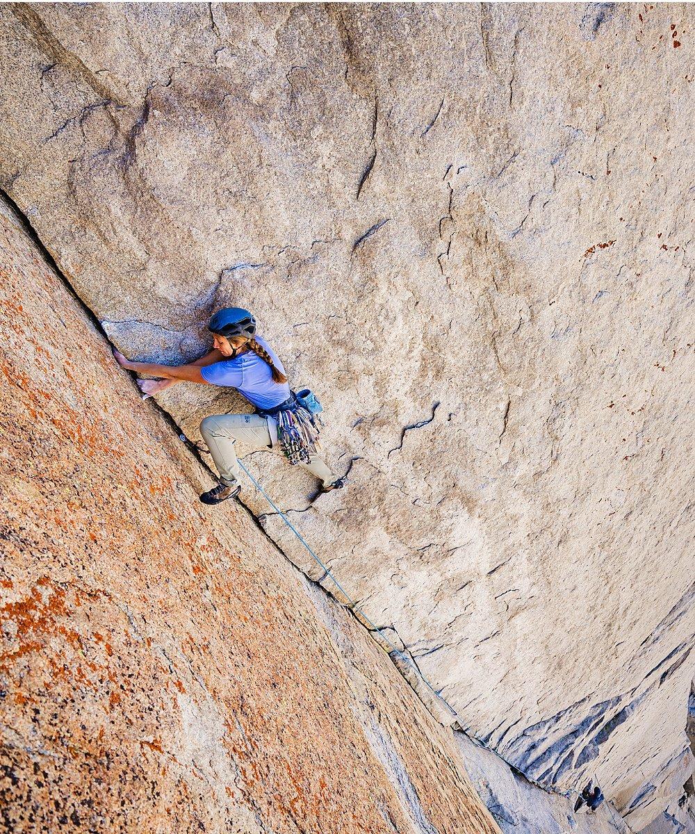 Climber Jen Poe ascends a steep granite wall on "The Incredible Hulk" towers in the Sierra Nevada, wearing a light blue Crater Lake™ short sleeve shirt, a helmet, and climbing gear with a rope trailing below her.