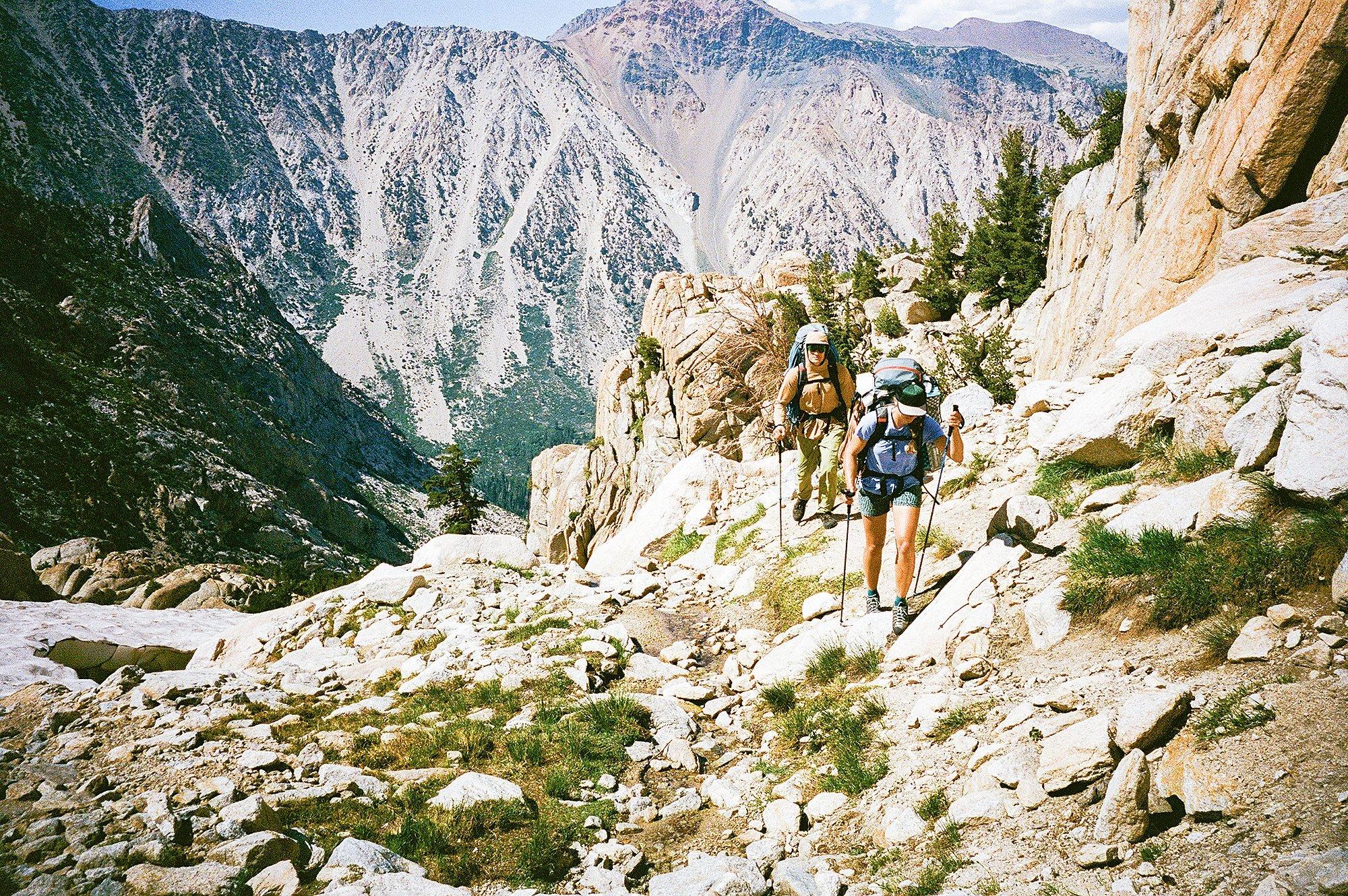 Jen and Casey, wearing large packs and using trekking poles make their way up a rocky mountain trail in the Sierra Nevada. The trail is surrounded by rugged granite terrain and scattered alpine vegetation, with steep cliffs and dramatic peaks rising in the background under a partly cloudy sky.