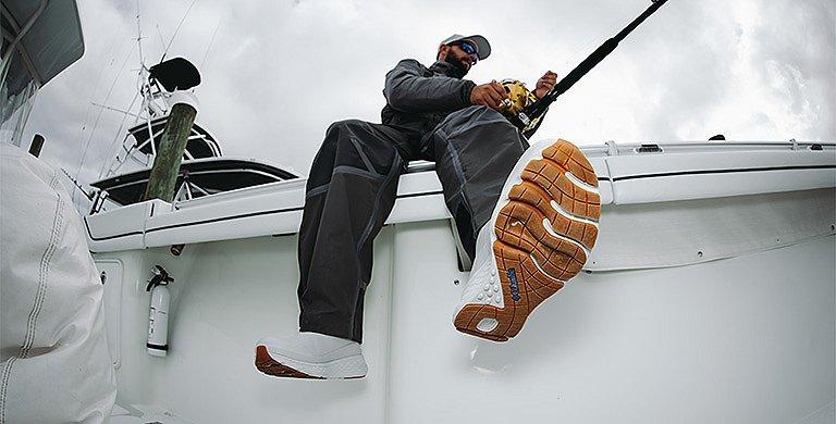 A man sits on the rail of a boat while reeling in his line; the grippy, textured soles of his PFG Pro Sport Boots are visible 