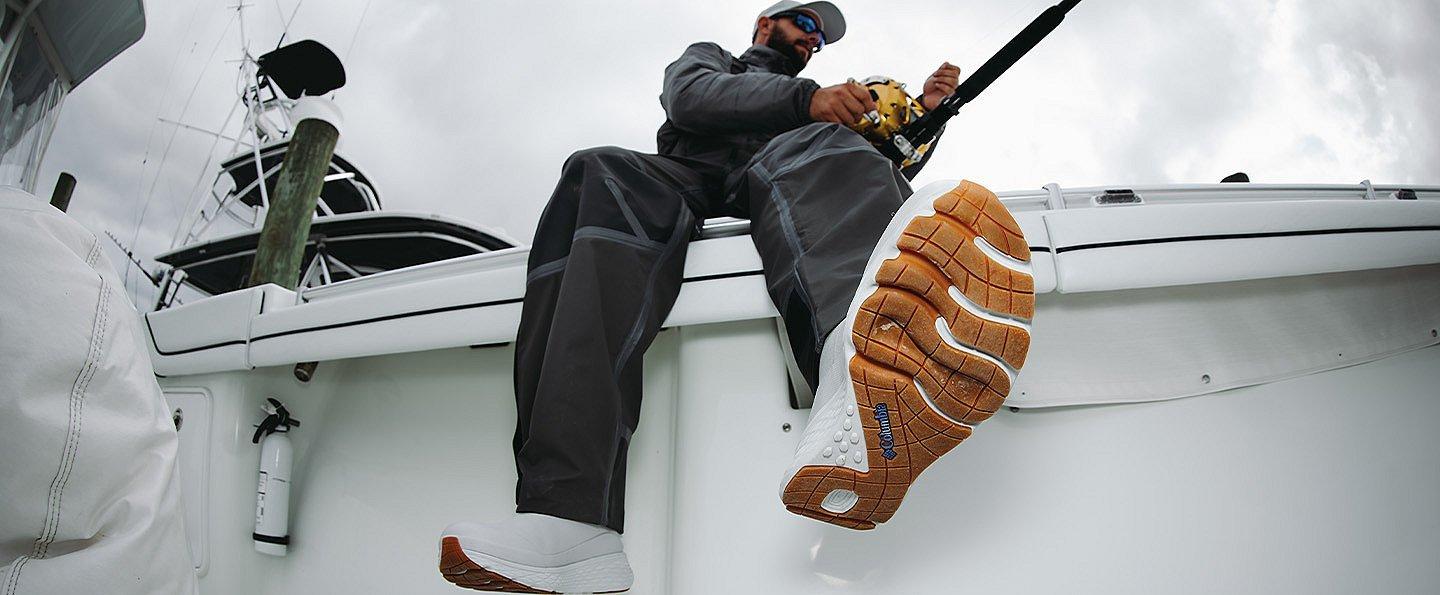 A man sits on the rail of a boat while reeling in his line; the grippy, textured soles of his PFG Pro Sport Boots are visible 