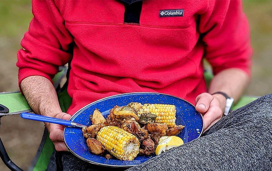 A man in a red Columbia Sportswear jacket sits in a camping chair outside holding a blue dish full of grilled food.