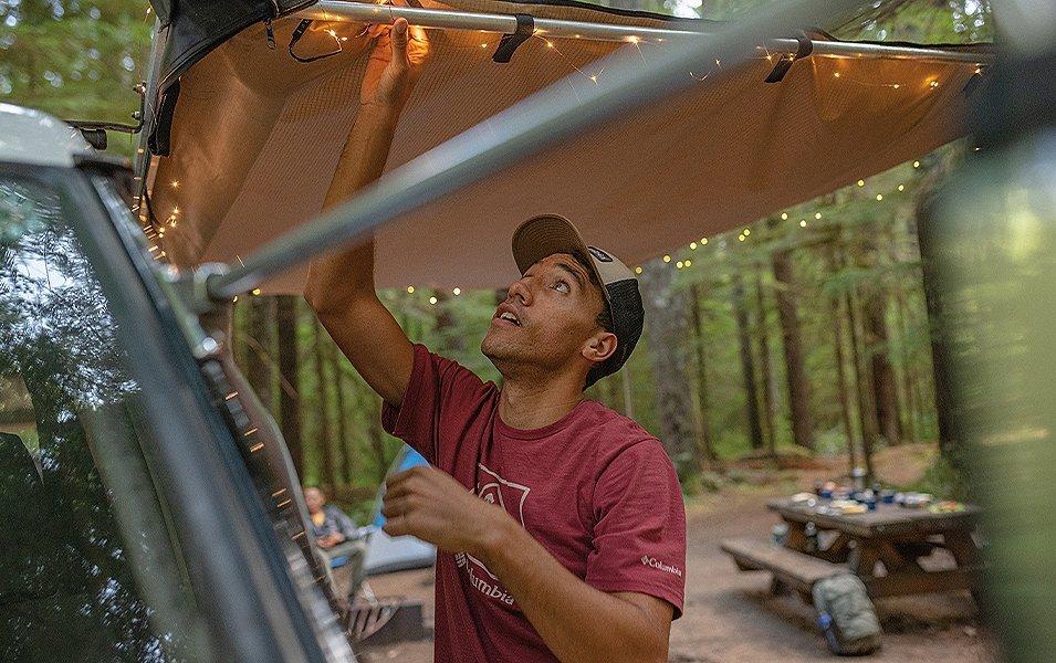 A man connects string lights to a van parked in front of a picnic table at a scenic campground.