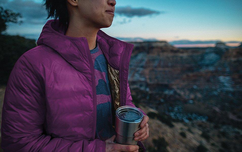 A woman wearing a purple Columbia Sportswear stands in the mountains holding a warm cup of coffee and looking out over a canyon.
