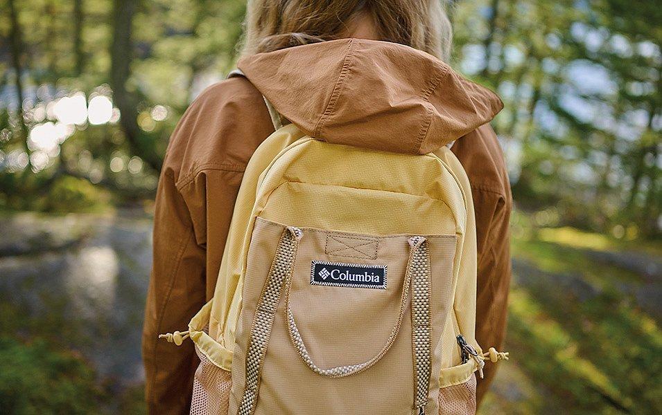 Above a tree-lined lake, a woman sports a Columbia backpack and rainshell. The gear’s muted earth tones blend into the alpine stillness