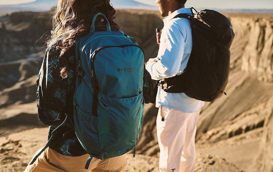A man and woman, packs on, stand on a desert plateau, surveying the sweeping desert landscape.