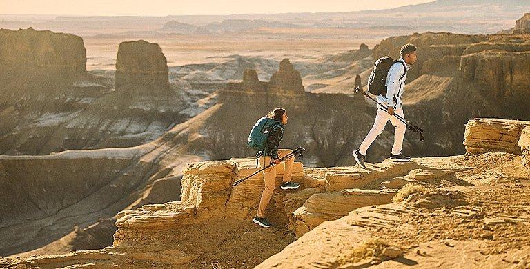 Two hikers in sun-protective gear hike above a vast desert canyon.