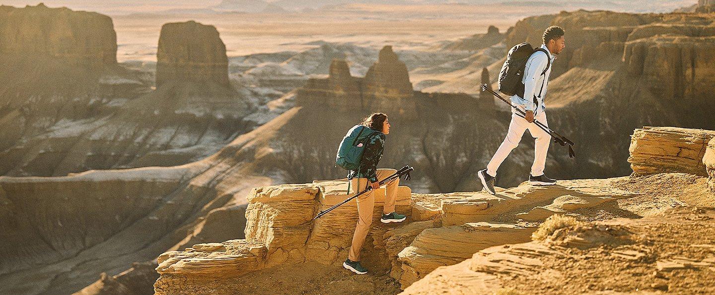 Two hikers in sun-protective gear hike above a vast desert canyon.