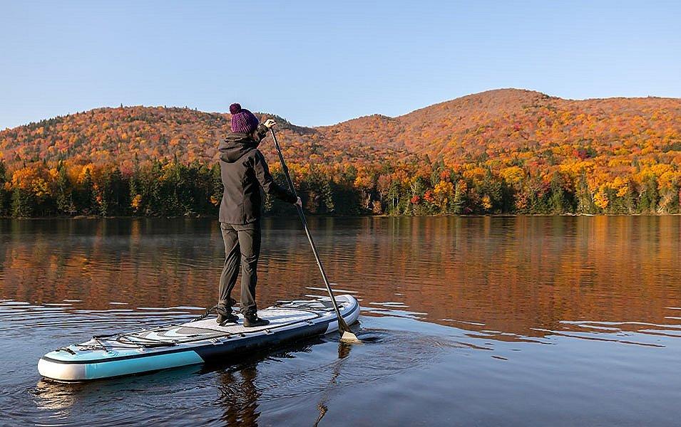 A woman wearing a hoodie and warm beanie paddles a stand-up paddleboard during a fall or winter adventure.