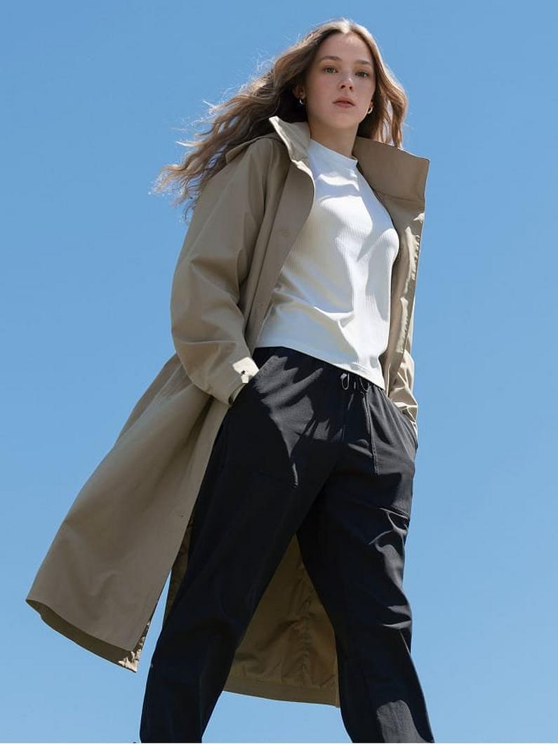 Woman wearing beige long coat over white hoodie and black jogger pants, standing outdoors.