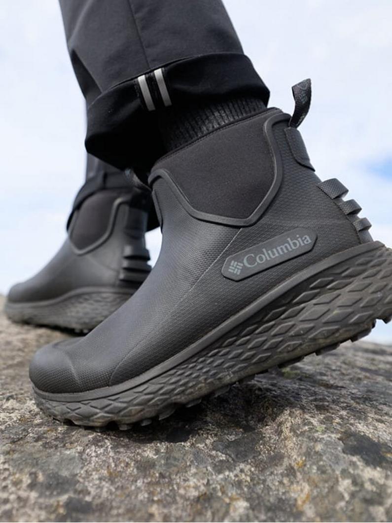 Person walking on rocky terrain wearing black slip-on ankle boots with textured rubber soles.