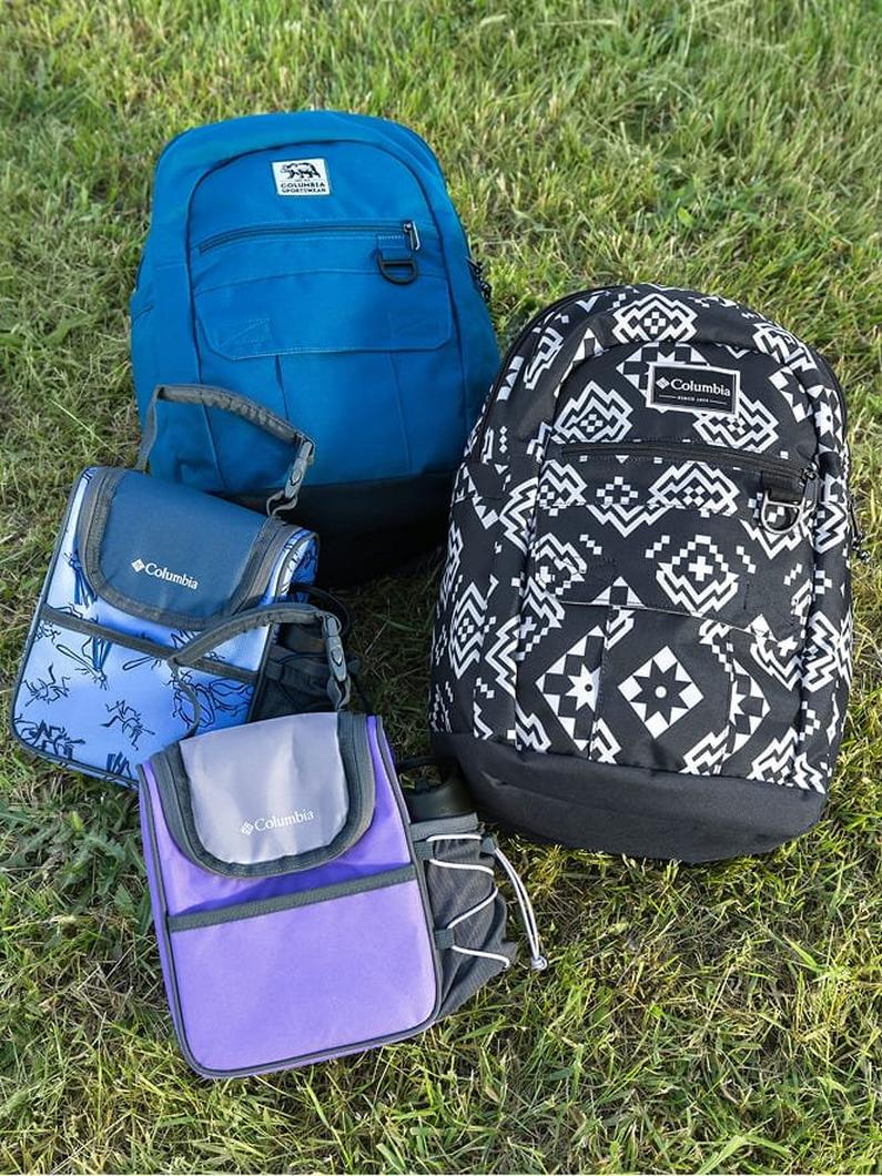 Collection of backpacks and lunch bags in blue, black geometric print, and purple displayed on grass.