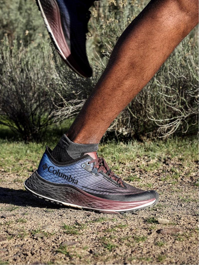 Close-up of person wearing blue and black athletic trail running shoe with gray sock on outdoor terrain.