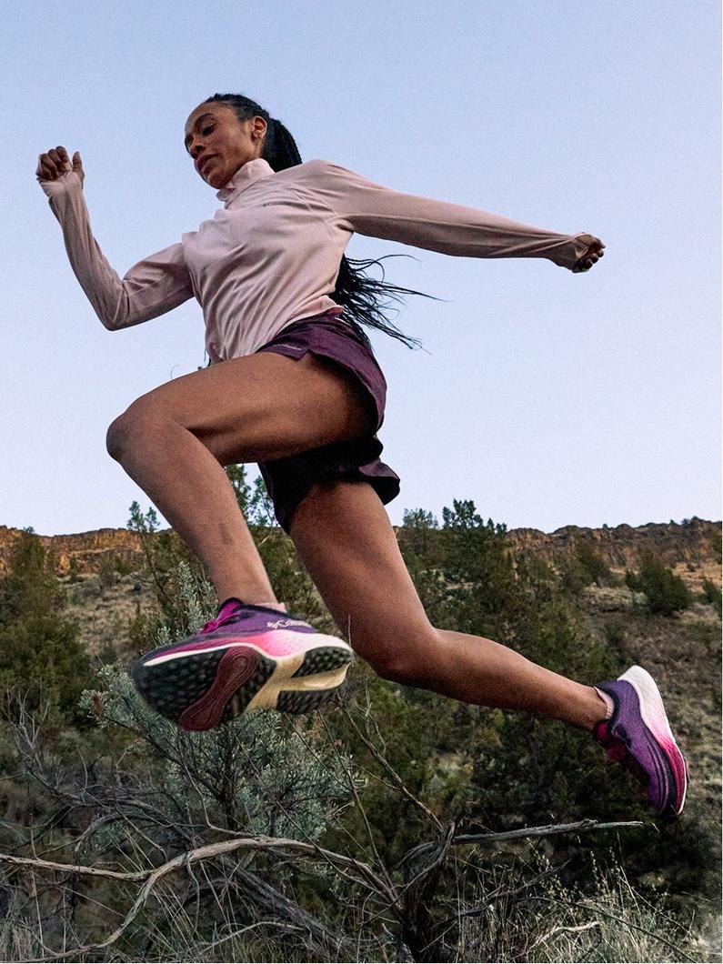Woman running outdoors in purple running shoes, pink long-sleeve top, and burgundy shorts on a trail.