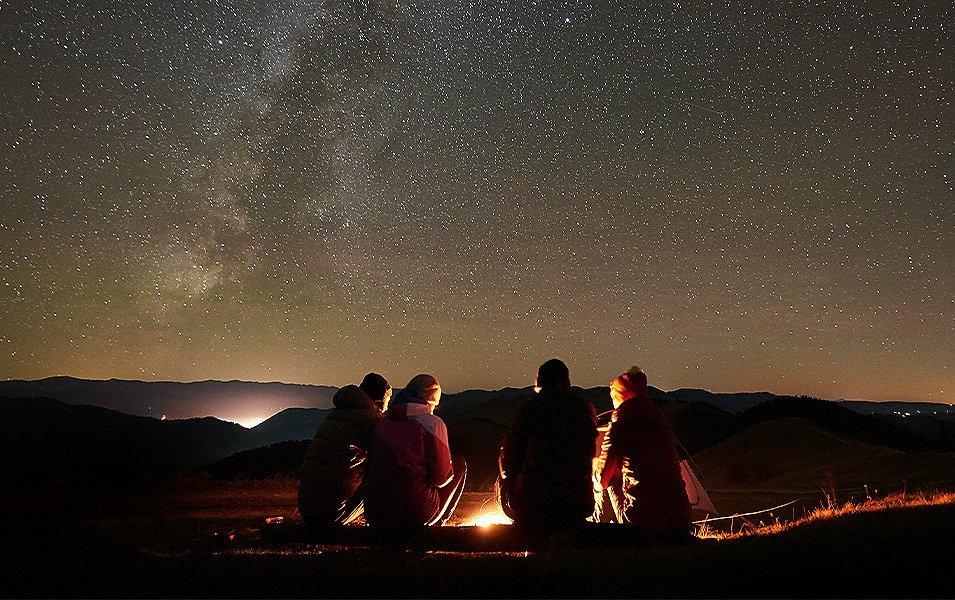 Four people are sitting on a fallen log, gazing into a glowing campfire. Behind them, stars light up the night sky.