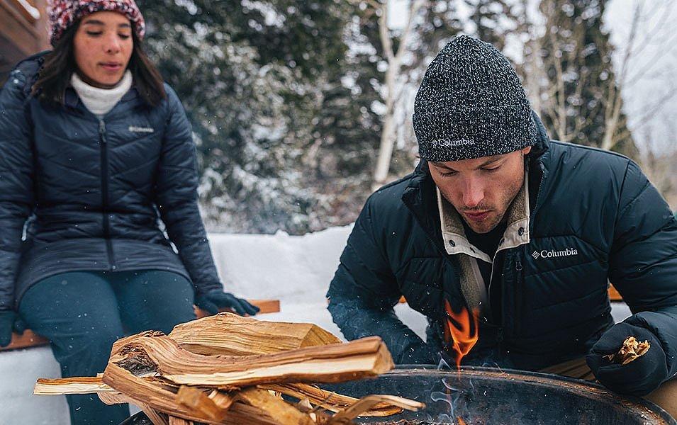 A woman stands behind a man as he tends to a campfire in a pretty forest setting.