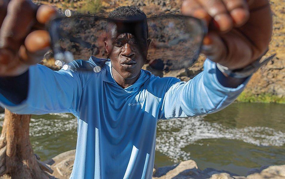 A man stands in front of a scenic river holding a pair of sunglasses to the camera.