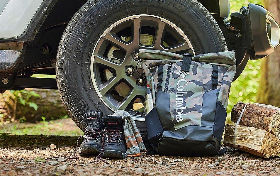 A Columbia Sportswear drybag sits next to a vehicle, alongside a pair of hiking boots and campfire wood